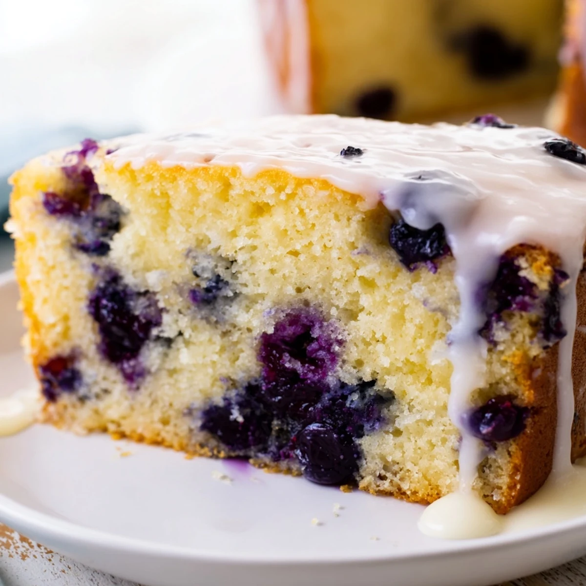 Golden slice of lemon blueberry cake showing purple berry flecks and drizzled with sweet lemon icing